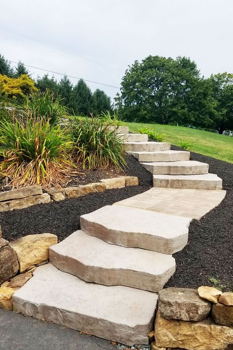 We designed and installed these stone steps that connected a driveway to an upper porch entrance area. Built into the existing slope of a hill, the landing areas between the steps incorporate smaller pavers with a stylish laying pattern. Steps and walkways are landscape elements that offer a great chance to express your unique style and enhance nature's beauty. Landscaping Stone Steps, Sloped Front Yard Steps, Landscape Ideas For Sloping Backyard, Outdoor Steps On A Slope Hill Landscaping, Landscaping Steps On A Hill, Waterfall Steps Front Porch, Landscape Steps On A Hill, Patio Built Into Hill, Outdoor Landscape Steps