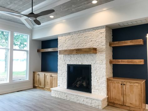 This custom floor to ceiling stone fireplace is accented with a hand hewn barn beam mantel and floating shelves. The accent wall color of a deep navy pops the white stack stone and maple woodwork right off the wall! Navy Wall White Fireplace, Fireplace Mantle Accent Wall, Accent Wall With Stone Fireplace, Navy Fireplace Wall Living Room, Accent Walls Next To Fireplace, Backside Of Fireplace Ideas, Navy And White Fireplace, White Fireplace With Accent Wall, White Fireplace Blue Wall