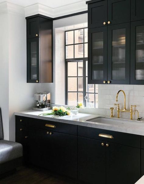 Black kitchen features glass reeded upper cabinets and black lower cabinets adorned with polished brass hardware paired with white marble countertops and a white subway tile backsplash. Black Kitchen, Black Cabinets, Counter Tops, A Kitchen, Kitchen Cabinets, Handles, Marble, Brass, Glass