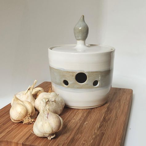 Handmade garlic keeper glazed in white with a gray band around the middle. The gray glaze has shades of brown and light blue.  Garlic crock is wheel thrown stoneware clay glazed with lead-free, food-safe, studio made glazes. Garlic crock has multiple holes to allow air flow while blocking the majority of light. Interior of lid is unglazed to absorb unwanted moisture. Depending on the size of your garlic, this keeper will easily hold 1-4 bulbs. Height: 3.5-inches (5-inches with handle) Width: 4.5-inches Weight: 1 lb, 4 oz Every effort to photograph color as accurately as possible is made, but I cannot guarantee that what you see is an exact representation due to the differences in monitors.  If you are uncertain, please contact me for additional photos. Garlic Keeper, Garlic Storage, Garlic Jar, Ice Blue Color, Kitchen Jars Storage, Hand Shapes, White Glaze, Jar Containers, Storage Jars