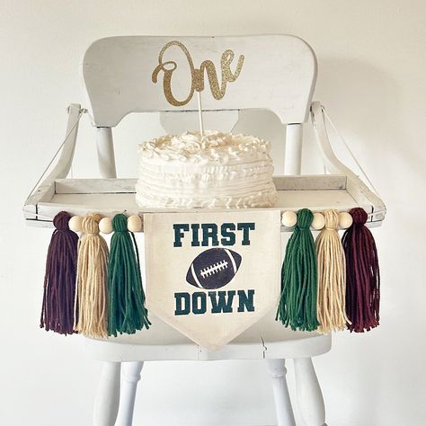 "For your football baby who is turning one, this 1st birthday highchair banner is the perfect sports flag to celebrate a milestone birthday. Green words \"First Down\" plus a brown football make this flag a classic for their first celebration.  Banner includes 6 tassels in dark green, tan and brown separated by wooden beads. Hang on a high chair or above the cake table and when the party is over hang on the wall as part of a room decoration.  This football cake smash sign makes an awesome photo prop and keepsake. DETAILS Full banner: This 1st birthday banner comes with the center canvas banner with 2 pieces of attached double sided tape, 6 acrylic yarn tassels, and 6 wooden beads. It arrives already strung on a string. Dowel is included if you'd like to hang it on the wall without the tass First Down Football Birthday Party Cake, First Down Bday Party, 1st Year Down Football Birthday Photoshoot, Football Birthday Decor, First Down 1st Birthday Party, First Down Cake Smash, Tailgate First Birthday Party, First Down Birthday Party Theme, 1st Touchdown Birthday