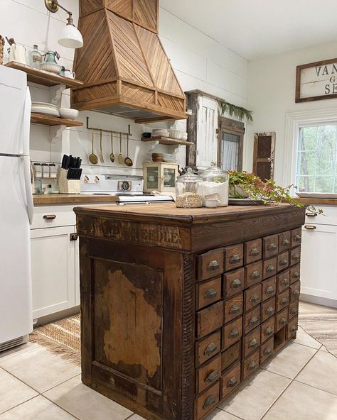 This kitchen with ivory cabinets boasts an antique wooden apothecary cabinet used as a rectangular kitchen island. The island stands on beige floor tiles in front of a stained wood range hood. Schoolhouse Kitchen, Rustic Kitchen Island Ideas, Apothecary Kitchen, Kitchen Island With Drawers, House Renos, Antique Kitchen Decor, Basement Redo, Kitchen Island Ideas, Rustic Kitchen Island