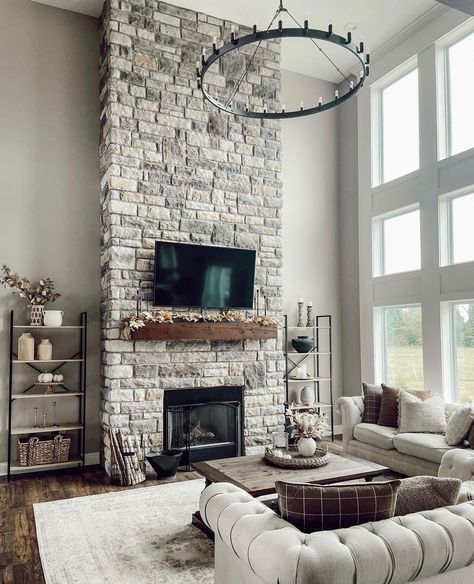 A flat panel TV is mounted to a gray stone two story fireplace above a stained wood mantel. The TV faces a light gray Chesterfield sofa and wood coffee table lit by a black wagon wheel chandelier. Stone Tile Fireplace, Shiplap Living Room, Fireplace With Tv, Stained Wood Beams, Farmhouse Fireplace Ideas, Stained Wood Trim, Grey Stone Fireplace, Two Story Fireplace, Stone Fireplace Wall