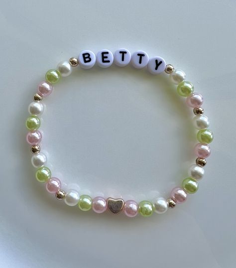 A white, pastel pink, and pastel green friendship bracelet, dotted with rose gold seed beads and a rose gold feature heart bead. The bracelet reads "Betty". Pastel Bead Bracelet, Bettys Garden Bracelet, Betty Friendship Bracelet, Betty Bracelet Taylor Swift, All Too Well Bracelet, Betty Bracelet, Bejeweled Bracelet, Concert Bracelets, Frendship Bracelets