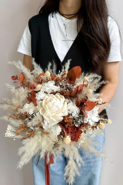 Your special day deserves a bouquet that reflects your free spirit and love for nature. This handcrafted boho bridal bouquet, bursting with warm terracotta, burnt orange, and sunshine yellow blooms, evokes the magic of autumn. Soft pampas grass adds a touch of texture, while luxurious cafe au lait roses whisper romance. Hand-tied with love, this bouquet is the perfect complement to your bohemian spirit and a forever memento of your rustic wedding celebration. Terracota Bridal Bouquet, Burnt Orange And Pampas Wedding Flowers, Dried Flower Arrangements Wedding, Burnt Orange Wedding Flowers, Dry Flower Bouquet, Boho Bridal Bouquet, Autumn Soft, Preserved Hydrangea, Preserved Eucalyptus