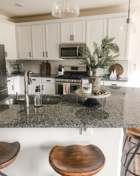 The dark gray speckled countertop of this angled island stands out against its light gray base. It includes a conveniently built-in sink and a cake stand with a candle, a bell, and a bottle holding an arrangement of evergreen branches. Gray Kitchen With Granite Countertops, White Cabinet Gray Granite Countertop, Kitchen With Dark Marble Countertops, Kitchen Decor With Granite Countertops, Black Speckled Countertops, Salt And Pepper Countertops Granite, Kitchen Ideas Florida, Dark Grey Countertops White Cabinets, Kitchen Gray Granite Countertops