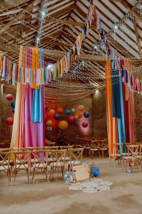 This shows a wedding ceremony set up in Octagon Barn, Norfolk. The theme is a vibrant colourful one, with colourful drapes and bunting hanging from the ceiling. Alongside the aisle are cross back wooden chairs. At the start of the aisle is a yellow welcome sign, surrounded by a jute rug, disco ball, flowers and lanterns. At the end of the aisle are wooden plinths with large floral installations on them, with hanging lanterns over the plinths. Floral Plinths, Colourful Bunting, Barn Wedding Ceremony, Barn Wedding Decorations, Crossback Chairs, Furniture Board, Disco Balls, Wooden Cross, Flower Fashion