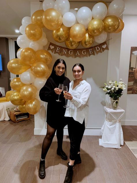 Two Indian women standing in front of decoration display made up or gold and white balloons and a banner that says engaged on it, cheersing with champagne glasses Simple Engagement Party Ideas At Home, Casual Engagement Party Decorations, Small Engagement Party Ideas Simple, Gold Engagement Party Decor, White And Gold Engagement Party, Engagement Party Decorations Indoor, At Home Engagement Party, Simple Engagement Party Decorations, Classy Engagement Party