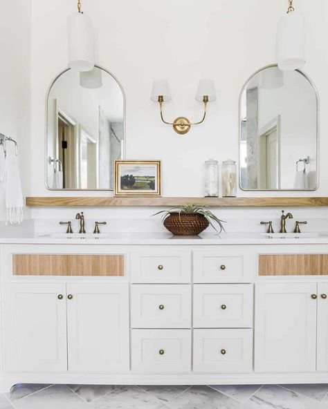 White and brass dual head sconce is flanked by silver frame arched bathroom mirrors mounted above a light wood floating shelf. A white dual sink vanity under the shelf is lit by white and brass pendant lights. Shelf Above Sink, Wood Mirror Bathroom, Bathroom Mirror With Shelf, Bathroom Mirror Storage, Rectangular Bathroom Mirror, Vanity Shelves, Bathroom Sconces, Wood Floating Shelves, Storage Mirror