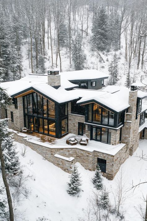 Aerial view of a snowed in modern mountain mansion with modern stone foundation with large porches and multi pitched second floor roof large windows. Indulge in luxurious mountain mansions that seamlessly blend natural beauty with luxurious living, offering a unique retreat away from the everyday hustle. Homes In Mountains, Modern Winter Cabin Exterior, Barndominium In The Woods, Modern Houses In The Woods, Mountain View House Plans, Luxury Mountain Homes Exterior, Mountain Chalet Exterior, Mansion In Woods, Modern Mountain Mansion