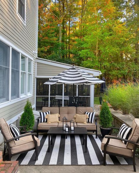 Tan patio seating rests upon a black and white striped outdoor rug accessorized by matching pillows and umbrella. Black table sits in the middle of the furniture showcasing black lanterns with candles. Potted shrubs complete the space. Black White And Green Patio Decor, Black And White Outdoor Rug Patio, Black And White Patio Decor, Potted Shrubs, Patio 2023, Striped Outdoor Rug, Polywood Outdoor Furniture, Black And White Patio, White Patio Furniture