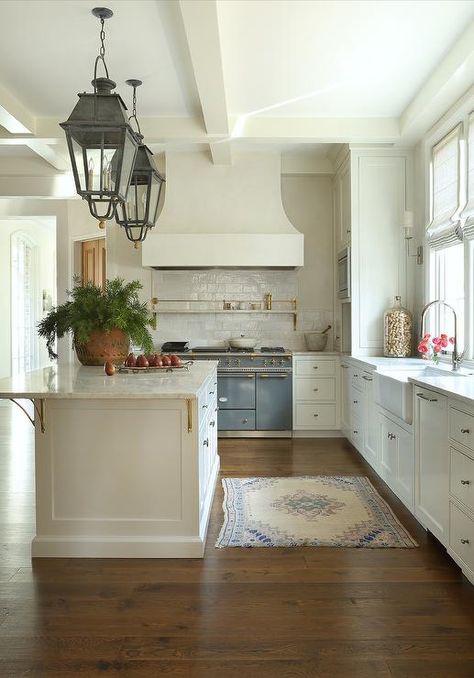 Large cream kitchen design features a blue French Lacanche range under a cream French hood on cream glazed backsplash tiles, black carriage lanterns over a cream kitchen island, a white farm sink with a polished nickel gooseneck faucet, and a blue and white vintage rug atop wooden floors. Cream And Blue Kitchen, White Farm Sink, White Kitchen Hood, French Stove, Cream Kitchen Cabinets, Blue Kitchen Island, Kitchen Island Tops, Cottage Kitchen Design, Mixing Metals