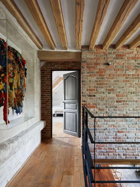 an upstairs hallway with wood plank floor and black metal railings, one cement wall with a fabric artwork, a brick wall with an dark colored open door leading into a bright bedroom, old wood beamed ceiling Minimalist Backdrop, Elle Decor Magazine, Brick Living Room, Bright Bedroom, Prefab Buildings, Fabric Artwork, Brick Interior, Modernist House, Horse Stable