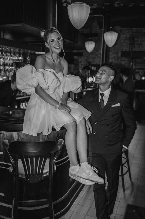 Groom gazes up at the bride sitting on the bar in a short wedding reception dress with puff sleeves nike sneakers and space bun hairstyle | bridal style | modern bride | short wedding dress | wedding hair | bridal shoes | flat wedding shoes | Lindsey Ria Photography Civil Wedding Dress With Sneakers, Short Wedding Dress And Sneakers, Wedding Dress With Sneakers Brides, Wedding Dress And Sneakers, Bride Sneakers Wedding, Wedding Dress With Sneakers, Wedding Sneakers For Bride, Reception Dress Short, Bride Sneakers