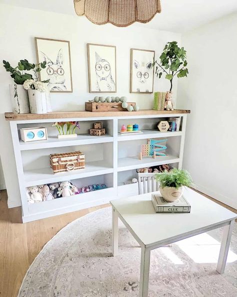 A trio of animal drawings in wood frames is hung above a white playroom shelving unit. Light from a nearby window illuminates the potted plants and toys accessorizing the shelves. A large wicker pendant light hangs over a miniature white table decorated with a small potted plant. Playroom Shelving, White Playroom, Green Playroom, Gray Playroom, White Small Table, Shiplap Feature Wall, Grey Painted Walls, Playroom Shelves, Wicker Pendant Light