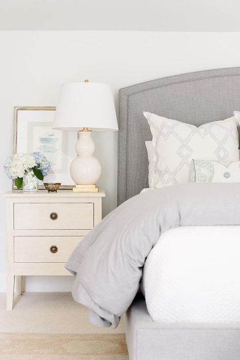 Lovely white and gray bedroom features an ivory wood French nightstand styled and topped with a white double gourd lamp. Night Stand Styling, French Nightstand, Grey Upholstered Bed, Gray Headboard, Nightstand Styling, Nightstand Design, Grey Headboard, Transitional Bedroom, Curved Headboard