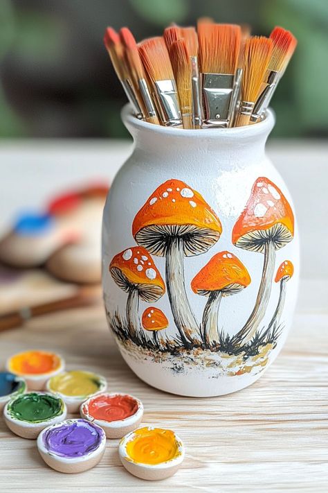 A painted pottery vase featuring orange mushrooms with brushes and colorful paint, ideal for cottagecore-themed decor. Painting Mushrooms Ideas, Mushroom Pottery Painting, Painted Pottery Ideas, Mushroom Painting Ideas, Mushroom Pottery, Painting Mushrooms, Pottery Painting Ideas Easy, Pottery Painting Ideas, Cottage Core Art