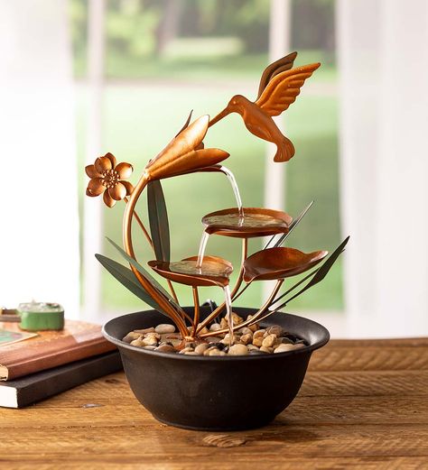 Place this charming Hummingbird Metal Fountain on your desk or in any room that could use the calming power of flowing water. Copper-colored metal recreates the scene of a hummingbird stopping for a snack at a flower. Green metal leaves complement the copper colored sculpture. The plastic basin holds the included smooth river rocks and conceals a quiet recirculating pump. Bring the sights and sounds of nature into your home with this delightful tabletop fountain. Recommended for indoor Decorating With Copper, Hummingbird Fountain, Copper Table Top, Indoor Tabletop Water Fountain, Table Fountain, Copper Garden, Fire Pits Outdoor, Hummingbird House, Fountain Ideas