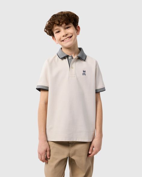 A smiling young boy with curly brown hair is wearing a beige KIDS SOUTHPORT PIQUE POLO SHIRT - B0K263B200 by Psycho Bunny made of Pima cotton piqué with grey collar and sleeve trim, and khaki pants. He stands against a plain light grey background. Polo Shirt Outfit Men, Polo Shirt Outfits, Polo Fashion, Shirt Outfit Men, Boys Tops, Polo Tees, Pique Polo Shirt, Swimwear Shorts, Boys Top