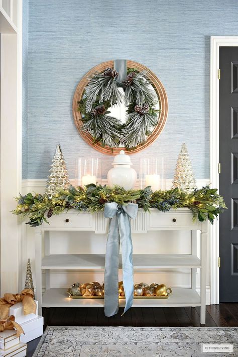 Console table styled for Christmas with a white ginger jar, glass hurricanes and mercury glass trees on either side. Lush greenery garland and frosted wreath. A classic grandmillenial home. Mercury Glass Trees, Christmas Entryway Decor, Glass Trees, Christmas Entryway, Chinoiserie Christmas, Blue Christmas Decor, Christmas Decor Inspiration, Christmas Tablescape, Christmas Time Is Here