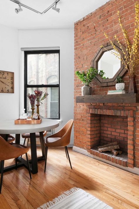 Corner dining space created in NYC apartment. Cement dining table is paired with leather dining chairs, highly durable and comfortable. An original brick fireplace sits as the backdrop with a dark wood floating mantle. We used organic accessories to style the space: rattan mirror, succulents, vase with tall branches, and a variety of glasses on the dining table. Natural Brick Fireplace, Floating Mantle Fireplace, Speakeasy Decor Bar, Fireplace Dark, Light Wood Dining Table, Floating Mantle, Speakeasy Decor, Neutral Wall Colors, Brick Living Room