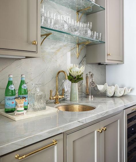 Vintage glass and brass shelves are mounted to a gold and gray marble slab backsplash between gray cabinets donning small brass knobs and over an antique brass vintage faucet accenting a brass pantry sink. Wet Bar Marble Backsplash, White Wet Bar, Shelves Over Kitchen Sink, Marble Slab Backsplash, Pantry Sink, White Brick Backsplash, Wet Bar Cabinets, Wet Bar Sink, Slab Backsplash