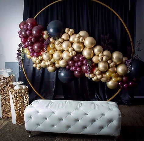 Set up a wondrous photo booth by using spectacular black polyester curtains hung over a backdrop stand placed behind a gold round metallic backdrop stand. Deck up the round arch stand with chrome gold, black, and burgundy latex balloons along with wine artificial silk flower garlands entwined with the balloons. Spread gold sequin runners upon display stands and position them beside the backdrop with white riser stands and white votive candle holders set on top for added finesse. Metallic Backdrop, Prom Photo Booth, Photo Booth Setup, Balloon Arch Frame, Event Decor Ideas, Party Decorations Table, Booth Setup, Balloons Flowers, Balloon Garland Diy