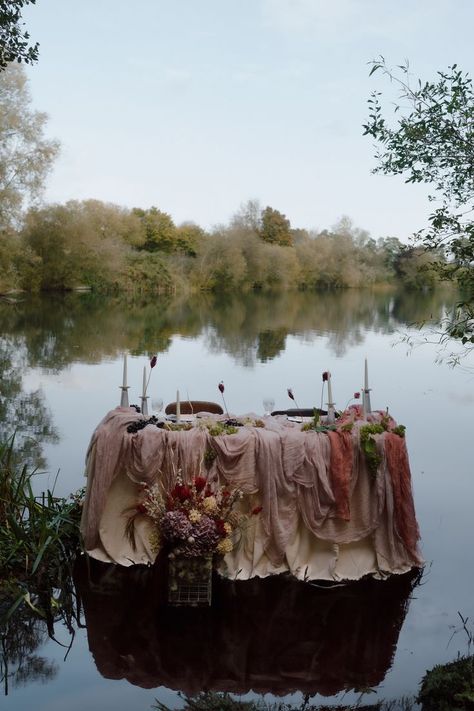 This shows a dining table in a lake. The table is covered in cream, violet and plum coloured drapes. On the tables are candlesticks, flowers, grapes and dinnerware. Behind the table are wooden chairs. In front of the dining table is a flower arrangement. Layered Drapes, Florist Studio, Event Display, Vendor Booth, Layered Fabric, Colour Texture, Tablecloth Fabric, Booth Ideas, Table Linen