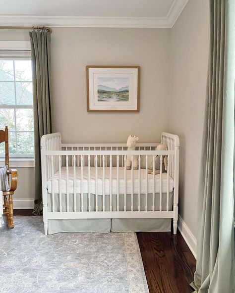 Sage green curtains frame light greige walls in this nature-inspired gender neutral nursery. A framed watercolor landscape hangs above a white wooden crib, where the sage green and white bedding coordinates with the room. A faded blue rug brushes the dark hardwood floors. Green And White Bedding, White Crib Nursery, Light Green Nursery, Blue Green Nursery, Green Nursery Boy, Sage Green Curtains, Dark Hardwood Floors, Wooden Crib, Greige Walls