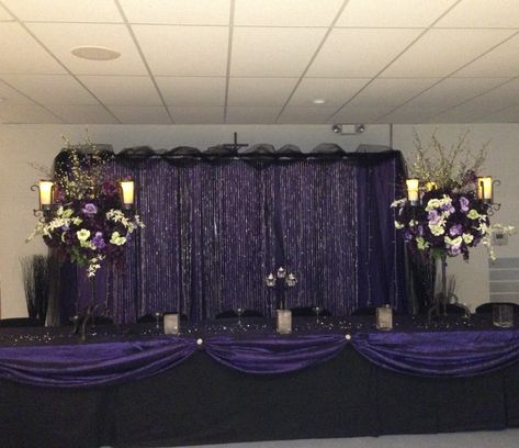 Elegant purple with gold accents backdrop with black tool across the top. Also has the head table decorated with purple swag with gold accents. Votive candles and purple/white flower arrangements too. Planning a wedding or event? Check out Cornerstone Interiors, Weddings by Cornerstone, and our venue near Great Bend, Kansas called Corinthians Hill. https://www.corinthianshilleventcenter.com/ https://www.weddingsbycornerstone.com/ https://www.interiorsbycornerstone.com/ Purple And Black Table Decorations, Black And Purple Wedding Reception, Black And Purple Wedding Theme, Wedding Entry Table, Quince Decorations Ideas, Quinceanera Venue, Great Bend Kansas, Purple Black Wedding, Purple Wedding Reception