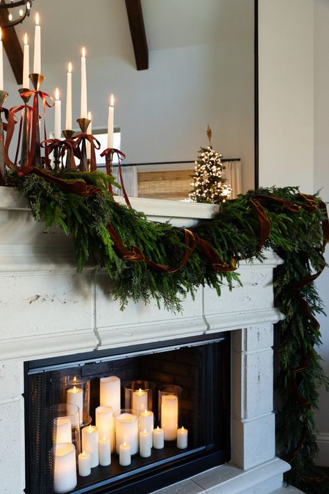 layered Christmas garland on fireplace mantle with brown velvet ribbon. Christmas bow trend using taper candles and brass candlesticks.  Lots of candles in the fireplace. Garland On Bookshelves, Christmas Garland On Fireplace, Candle Sticks On Mantle, Garland On Fireplace, Velvet Ribbon Christmas, Living Room Decor Ideas Cozy, Room Decor Ideas Cozy, Cozy Living Room Decor Ideas, Garland Mantle