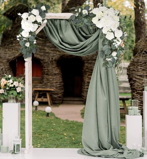 White wood rectangle wedding arch  with white rose and eucalyptus flower swag on each top corner. Eucalyptus color fabric draped from top middle to top right and flowing down right side of arch. Eucalyptus Ceremony Arch, Eucalyptus And Cream Wedding, Arch Spray Wedding, Olive Green Wedding Arch, Outside Gazebo Wedding Ideas, Simple Green And White Wedding Decor, Eucalyptus Wedding Backdrop, Swags For Wedding Arch, Gazebo Decorating Ideas Wedding Simple