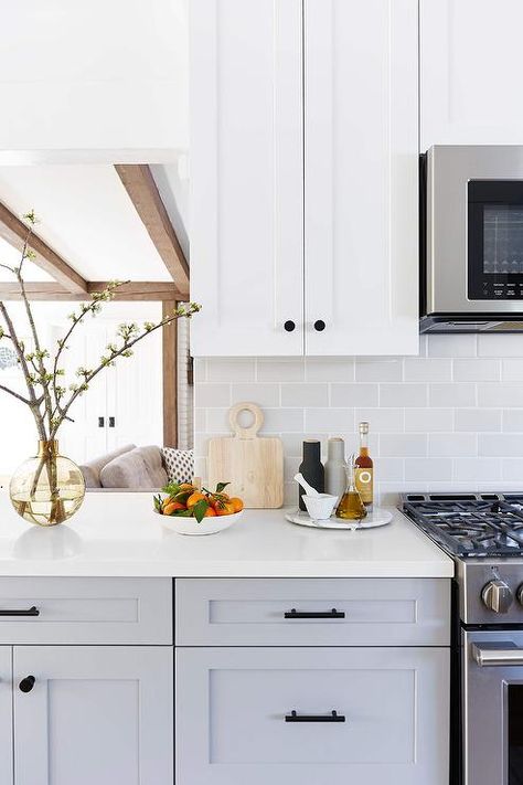 White shaker upper kitchen cabinets are mounted against light gray subway backsplash tiles and over gray shaker lower cabinets donning matte black hardware and a white quartz countertop located under a pass-through. White Upper Cabinets, Light Grey Kitchen Cabinets, Light Grey Kitchens, Upper Kitchen Cabinets, Gray And White Kitchen, Shaker Kitchen Cabinets, White Quartz Countertop, Grey Kitchen Cabinets, Shaker Kitchen