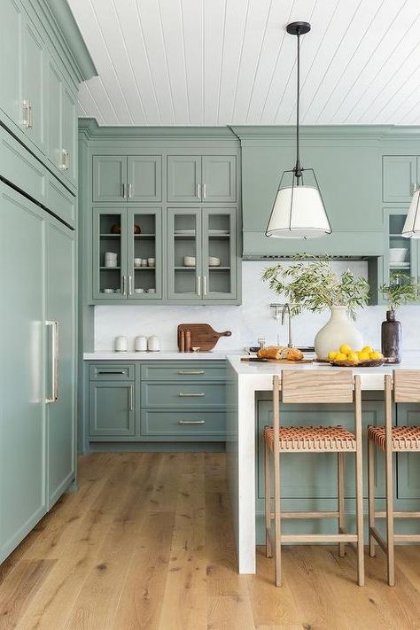 Kitchen features a plank ceiling over sage green cabinets with brass pulls, sage green wooden kitchen hood over white marble slab backsplash, polished nickel swing arm pot filler, kitchen island with white marble waterfall, brown oak and orange leather woven counter stools, illuminated by black and white cage drum lanterns. Taupe Kitchen Cabinets, Taupe Kitchen, Green Kitchen Designs, Sage Green Kitchen, Painted Kitchen Cabinets Colors, Green Kitchen Cabinets, Cabinet Paint Colors, Green Cabinets, Kitchen Cabinet Colors