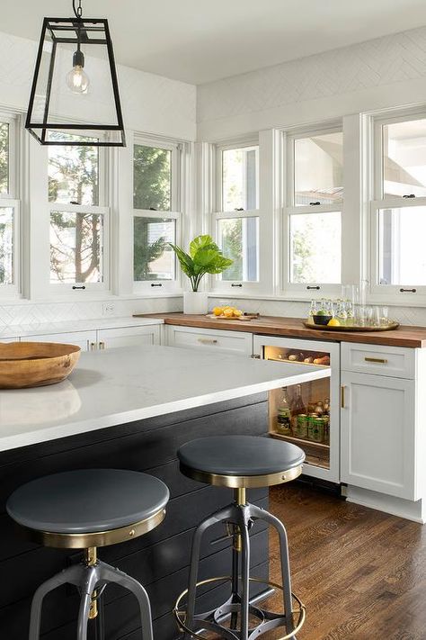 Beneath a row of windows, a butcher block countertop accents white shaker cabinets fitted on either side of a glass front beverage fridge, while Restoration Hardware 1940s Vintage Toledo Barstools sit at a black shiplap island topped with a white quartz countertop lit by filament pendants. Butcher Block Countertops With Quartz Island, Butcher Block And Quartz Countertops Kitchen, Butcher Block Quartz Countertops, Black Shiplap Island, Quartz And Butcher Block Countertops, Butcher Block And Quartz Countertops, Butcher Block Countertops White Cabinets, White Shaker Cabinet Kitchen, Black And White Farmhouse Kitchen