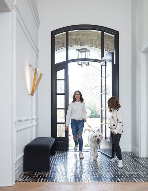 The arched front door’s sidelights and transom window flood this home’s double-height entry with natural light. A minimalist sconce adds a soft glow to the black and white foyer. | Photographer: Kim Jeffery | Designer: Shirley Meisels, MHouse White Foyer, Black Powder Room, Arched Front Door, Noguchi Coffee Table, Purple Sofa, Wooden Dresser, Transom Windows, Concept Home, Black Tiles