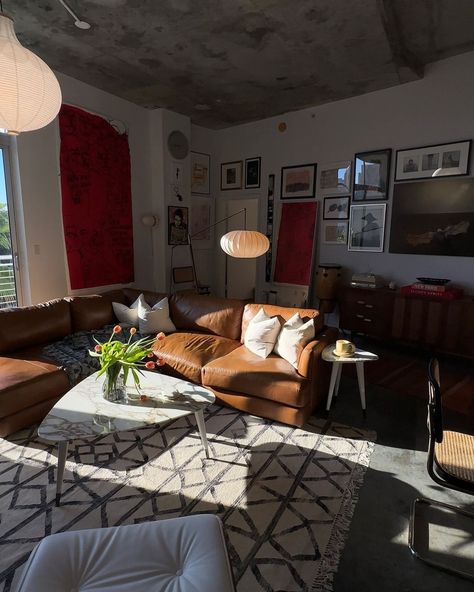 Photo of a modern living room with a brown leather couch, a white coffee table and red walls. Sunlight is coming in from large windows. The ceiling is concrete. There is a gallery wall with framed pictures. Warm lighting gives the room a moody, modern style. A white rug and black furniture complete the look. Brown Leather Couch Aesthetic, Leather Couch Apartment, Leather Sofa Living, Brown Leather Couch, Mid Century Modern Living Room, Leather Couch, Black Furniture, Red Walls, Living Room Inspo