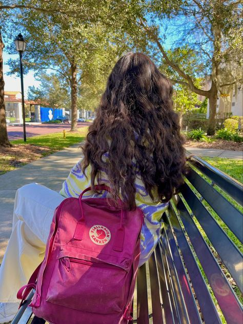 Someone sitting on a black bench with a deep pink kanken backpack. The person has long, dark, and wavy hair. They are wearing a green, purple, and white sweater with white pants. There are green trees and black lampposts in the background. Fjallraven Kanken Outfit, Fjallraven Kanken Backpack Aesthetic, Kanken Backpack Outfit, Kanken Backpack Aesthetic, Aesthetic Kanken, Kanken Backpack Mini, Backpack Kanken, Backpack Aesthetic, Backpack Fjallraven