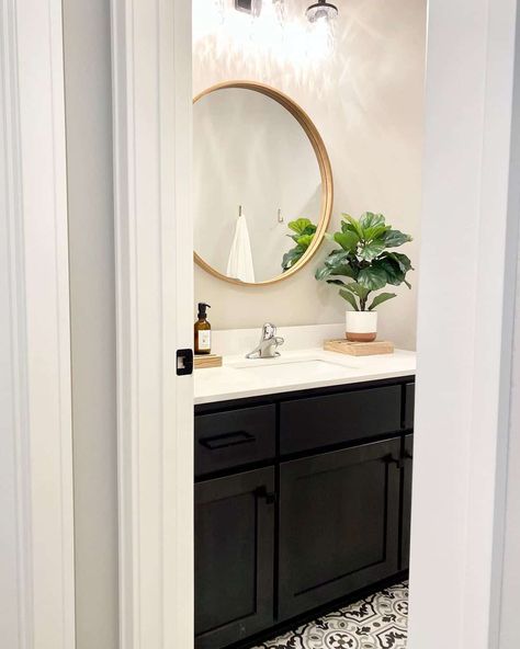 This bright bathroom uses vanity lighting to showcase the bathroom mirror. The circular mirror is complete with a wooden frame and anchored to an off-white wall. Black, white, and gray floor tile contrasts with the black vanity. Black Vanity Bathroom Floor Ideas, Black And White Flooring, Blue Kitchen Designs, Gray Floor, Black Vanity Bathroom, Grey Floor Tiles, Off White Walls, Bright Bathroom, White Tile Floor