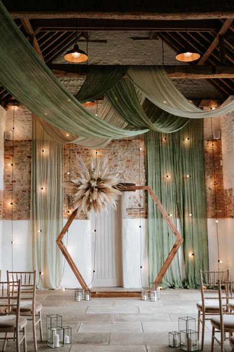 This shows a ceremony area at Curds Hall Barn, Norfolk. The focus is on a hexagon backdrop at the end of the aisle with a dried flower arrangement on the top left corner. At the base are some black box lanterns with candles. Behind and above the hexagon are green ceiling drapes paired with festoon wall lighting. Rustic Wedding Set Up Receptions, Sage Wedding Backdrop, Wedding Backdrop Drapes, Barn Draping Wedding, Modern Barn Wedding Decor, Green Wedding Backdrop, Hexagon Backdrop, Activation Booth, Unique Backdrops