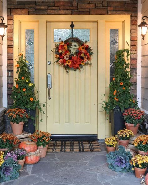 A creamy yellow door sets the stage for a warm and inviting entrance, while the plaid door mat adds a touch of charm. Standing tall on either side of the door, two potted trees lend a touch of grace to the scene. Surrounding the area, a delightful collection of potted mums in shades of yellow, gold, and orange, along with pumpkins of all sizes and textures and purple kale creates a captivating display. Learn how to decorate your fall porch this season on a budget Diy Outdoor Fall Decor, Floor Lanterns, Evergreen House, Potted Mums, Outdoor Fall Decor Ideas, Fall Front Door Decor, Outdoor Fall Decor, Wood Exterior Door, Door Paint Colors