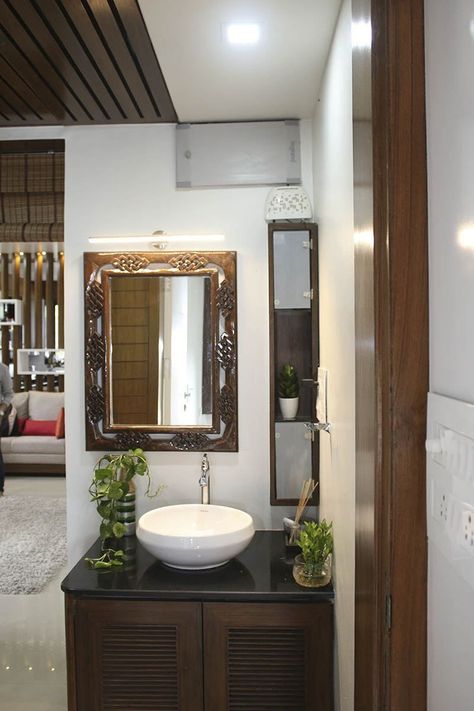 Common Basin Area incorporating Table Top Wash Basin on Black Granite Counter with Indoor Plants, Wooden Cabinet having Louvered Doors, Wall Mounted Rectangular Mirror with Wooden Frame highlighted with Wall ✨ Light and Vertical Wooden Cabinet with Glass Doors and Open Display Cabinet - GharPedia Table Top Sink Design, Granite Washbasin Counter, Basin With Cabinets, Basin Cabinet Design Modern, Pvc Farnichar, Granite Table Top Wash Basin Ideas, Wash Basin Ideas In Living Room Indian, Wash Basin Cabinet For Living Room, Table Top Basin Counter Design