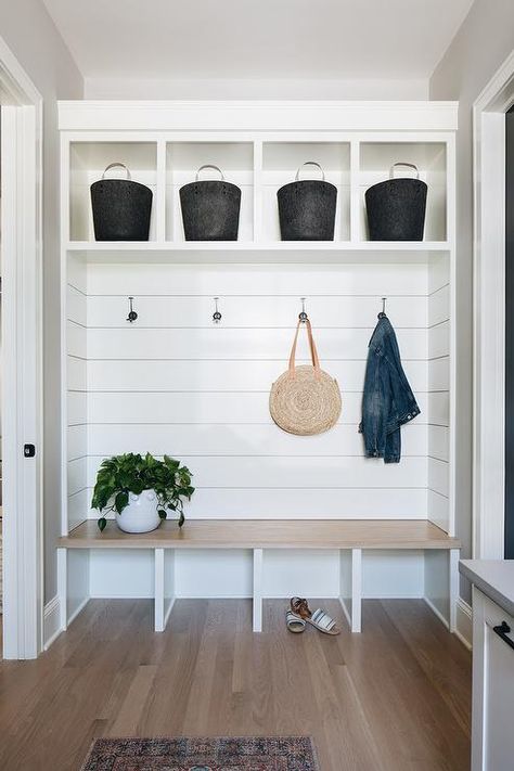 This welcoming mudroom features a light wooden bench fixed against a shiplap backsplash beneath coat hooks mounted under white cubbies holding black baskets. Shiplap Nook Wall, Built In Bench Open Underneath, Custom Entry Bench, Kitchen Nook Mudroom, Open Bench Seating, Entry Way Built In Bench With Storage, White Oak Mudroom Bench, Mudroom Seating Ideas, Entry Way Bench Built In