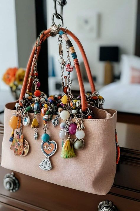 This image displays a light pink canvas handbag, with the handles wrapped in multicolored beaded decorations. Hanging from the handles are an assortment of charms, including colorful beads, tassels, and a heart-shaped pendant with the initials "CR." The combination of various shapes, textures, and colors gives the bag a playful, handcrafted feel, perfect for someone looking to make a bold, personalized fashion statement. Handbag Charms Diy, How To Make Purse Charms, Diy Purse Charms, Charms For Bags, Diy Bag Charm Ideas, Purse Charms Diy How To Make, Bag Charms Diy, Diy Bag Charm, Purse Charms Diy