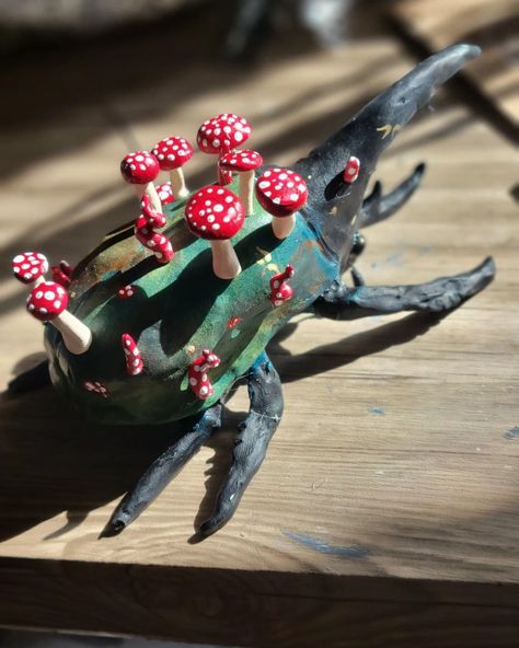 Large beetle with black head, black legs, and green body, facing away from camera, topped with red and white spotted mushrooms. Small gold flecks are scattered across the beetles body, some in the shapes of fae folk wings Polymer Clay Beetle, Ceramic Beetle, Bug Ceramic, Clay Beetle, Weird Sculptures, Clay Bugs, Hercules Beetle, Easy Clay Sculptures, Clay Inspo