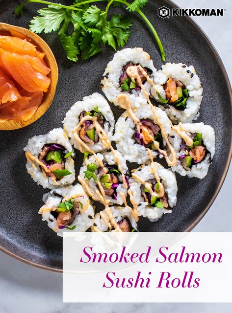 Image shows a dark gray plate, filling almost the entire frame, loaded with 10 sushi bites filled with smoked salmon, cucumber, purple cabbage, and cilantro. The rolls are drizzled with sriracha mayo. Also on the plate is a little wooden bowl with more smoked salmon, and the plate is garnished with a sprig of cilantro. The Kikkoman logo is at the top right of the image and the text overlay reads "Smoked Salmon Sushi Rolls" in a purple font on a transparent white background. Salmon Cream Cheese Sushi Roll, Sushi With Smoked Salmon, Smoked Salmon Sushi Recipes, Cooked Salmon Sushi Rolls, At Home Sushi Recipes, Smoked Salmon Sushi Roll, Diy Sushi Rolls, Smoked Salmon Rolls, Salmon Sushi Recipes