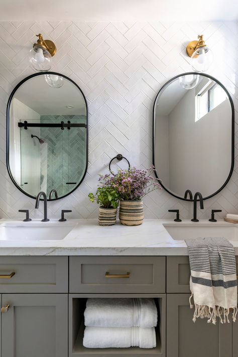 We are daydreaming of this perfect bathroom renovation. The muted gray color of the custom cabinetry with white countertops creates a lovely contrast with the black plumbing fixtures. Adding to the design, we incorporated two mirrors with an organic shape and a black edge detail, harmonizing with the plumbing. To complete the look, we decided to place contrasting brass sconces for both mirrors. We believe the result of this bathroom is truly charming! Farmhouse Bathroom With Black Fixtures, Bathroom Brass And Black Fixtures, Bathrooms With Brass Fixtures, Black And White Bathroom Brass Fixtures, Powder Room Ideas Grey Vanity, Gray Black White Bathroom, Black Plumbing Fixtures Bathroom, Different Metals In Bathroom, Farmhouse Plumbing Fixtures