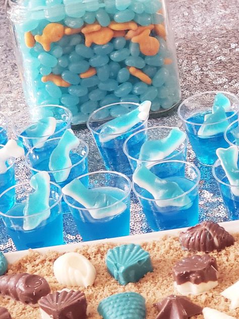 Blue jello in plastic shot cups with gummy shark on top.   In the jar behind, blue jelly beans with goldfish crackers. Blue Themed Food Platter, Blue Party Foods, Gummy Shark, Aquarium Birthday, Cup Desserts, Neverland Party, Halloween Jello Shots, Surf Birthday Party, Surf Birthday