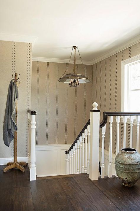 Beige rivet wallpaper complements white wainscoting and lines a white staircase accented with a dark stained wood handrail lit by a vintage lantern. Cottage Staircase, Staircase Wall Design, Staircase Wallpaper, Wallpaper Staircase, Handrail Lighting, 2025 Design, Transitional Staircase, Paper Weave, White Staircase