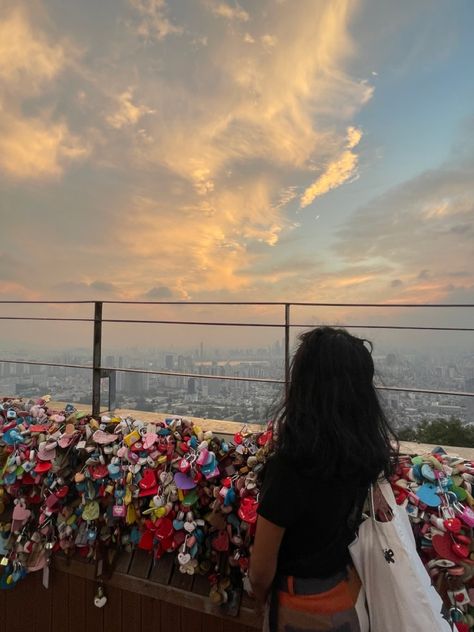 woman overlooking sunset, back facing towards the camera. She is wear a black shirt and patchwork pants. She is facing a railing covered in locks. South Korea Study Abroad, N Seoul Tower Aesthetic, Seoul In Summer, Summer In Korea Aesthetic, Seoul Instagram Story, South Korea Trip Aesthetic, Study Abroad Japan, Study Abroad Aesthetic Korea, Seoul Instagram Pictures